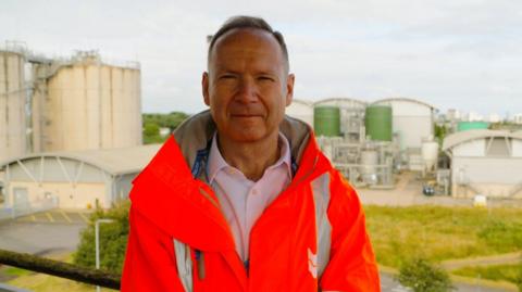 Roch Cheroux, a man with short greying hair, wearing an orange fluorescent jacket in front of an industrial backdrop.