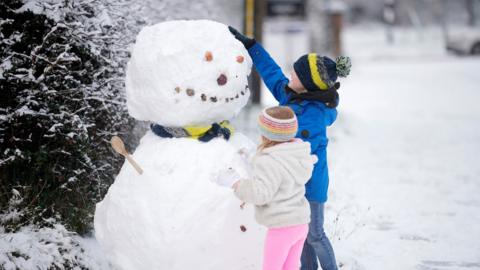 A young boy and girl, wearing blue and pink clothes, patting down snow on a snowman bigger than them
