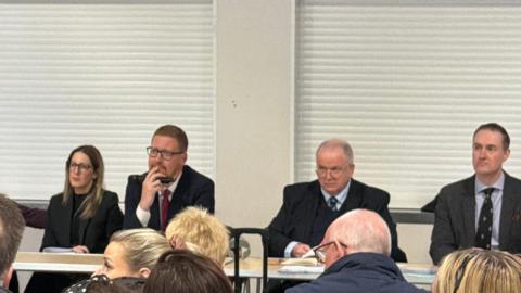 Community meeting attended by Jonathan Brash, Hartlepool Councillors Melanie Morley and Moss Boddy and Stockton North MP Chris McDonald. All four of these are pictured wearing dark black/grey suits. They are sat at a long table in front of an audience. A few members of the audience are in the image, but only the backs of their heads are visible.