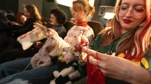 Women sit in a cinema and knit and crochet. 