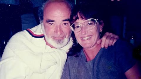 Eli Getreu is seen here with his wife Barbro Gentele. They are sitting close together and Mr Getreu has his arm around Ms Gentele. He is wearing a white top with a white jumper around his shoulders. She is wearing glasses and a blue dress and a watch.