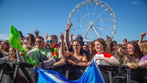 A crowd of people wearing summer clothes stand behind a barricade watching a performance. Two people at the front hold a Scotland flag. There is a ferris wheel behind them.