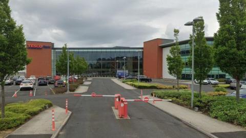 Tesco Bank's offices at Quorum Business Park are within a multi-storey glass building surrounded by a car park and trees. The bank's blue and red logo is placed between pillars near the top of the building.