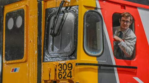 Brad Wright leans out of the Clacton Express train's driver side window and smiles at the camera. He wears a green and white striped shirt with green tie and glasses. The train has windows on the front and numbers. It is partially painted yellow with some grey, white and red.