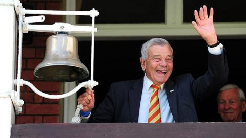 Retired international cricket umpire Dickie Bird rings the five minute bell prior to the start of play during day one of the 1st Investec Test match between England and New Zealand at Lord's Cricket Ground. Bird wears a dark jacket and a red and yellow striped tie and is waving with his left hand as his right pulls the bell.