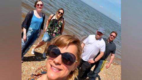 A selfie style photo of five adults standing on a beach. 