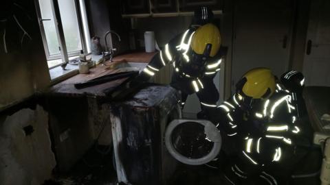 Two firefighters in protective clothing and wearing yellow helmets inspect a burnt tumble dryer following a blaze which has caused significant fire damage to the room it is in.