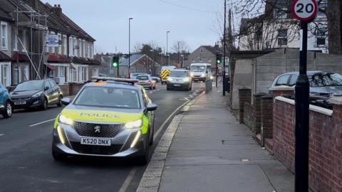 A police car is seen parked outside a property on Bensham Lane, a residential road. Cars at a traffic light junction can be seen in the background.