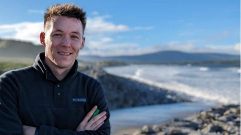 Nick Sheridan smiles at the camera with his arms folded. He has brown hair and wears a dark fleece. He is outside near a coastal scene and in the background there is a rocky beach with gentle blue waves. It is a sunny day.