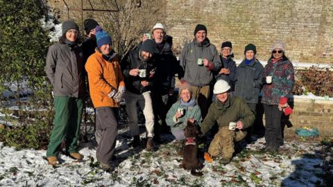 Twelve volunteers and a dog gather together in front of a wall in an ornamental garden. Six of them holding a cup of tea.  