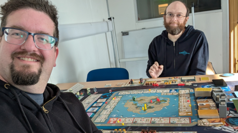 Rikh on the left is taking a selfie with Stuart. They are sitting at a table with a complicated board game at play in front of them