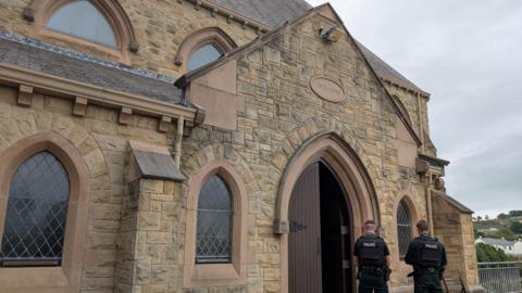 St Patrick’s Church in Downpatrick. It is an old brown building with a large brown door. Two PSNI officers are stood in front of the door with the backs to the camera. The sky is overcast. 