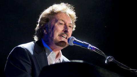 Supertramp founder Rick Davies wearing a suit and singing into a microphone. He has medium-length, light brown hair and a short beard and moustache. He is sat at a piano and the background is entirely black. 