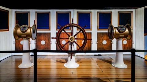 A wooden ship's wheel and compasses on display on a fake wooden floor with a fake ship display behind it.
