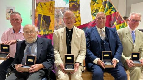 Five men in suits of different colours hold small boxes with medals in. There is a pink, green and yellow tapestry behind them. 