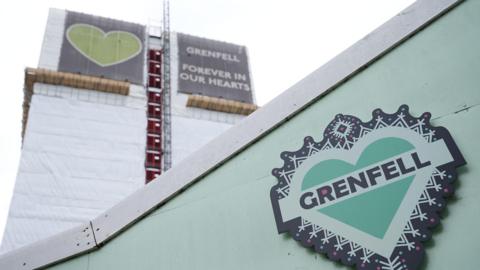 A view of a green fence, which has green and black artwork on it featuring a heart bordered by a line of stick figures and the word 'Grenfell' in the middle. Behind it, the shell of Grenfell Tower stands covered by tarpaulin and the 'Grenfell, forever in our hearts' signage at the top