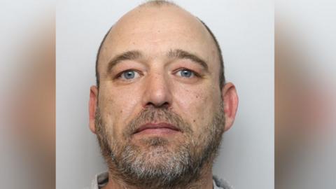 A mug-shot of Michael Sergio Smith, looking straight at the camera, he has blue eyes, short hair, and a beard. 