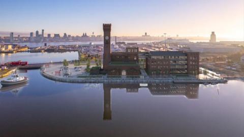 An artist's impression of the regenerated tower shows the original, brick-built two-storey pump room and chimney, with a new building rising up to about the same height as the pump house. The building stands on the front of a dock, with a car park next to it. The Liverpool waterfront can be seen across the Mersey in the background