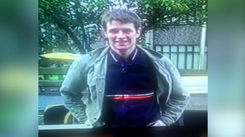 A young man with brown hair stands with his hands on his hips and smiles. He is wearing a khaki jacket over a blue fleece with a white and red stripe across the middle. Underneath he is wearing a navy polo shirt. He is in front of a street with a pavement, grey building with windows and trees.
