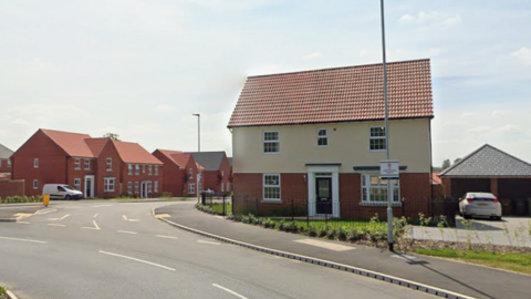 The picture is taken at the front of a housing estate which has one house on the right hand side and loads of red bricked houses on the left. A road is leading down to the housing estate. 