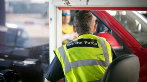 Man wearing hi vis jacket reading 'Border Force' sits in a booth while a car drives past