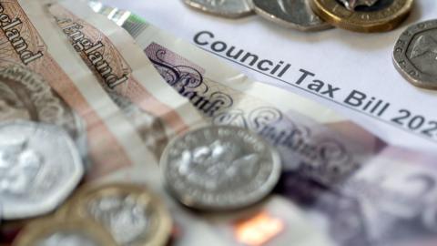 A council tax bill covered in money. The bill has a £20 note, two £10 notes and lots of pound coins, 50p and 20p coins on top of it.