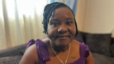 Christiana Hansen is looking straight into the camera, she is sitting on a sofa in her living room. She is wearing a purple dress and a gold chain.