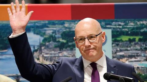 John Swinney, in blue suit, white shirt and purple tie, smiles and waves as he stands on a podium with a photo of Perth behind him.