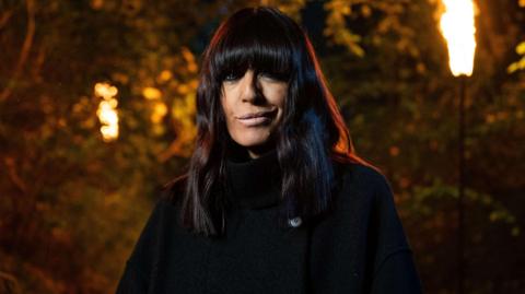 Claudia Winkleman dressed in a black cloak outside with fire on large lamps burning behind her. 