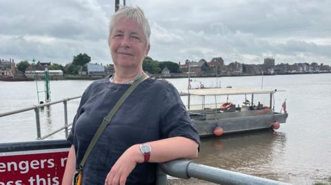 A woman standing in front of a river. Behind her is a grey boat. She is leaning on a metal railing. She is wearing a black top and has short grey hair. 