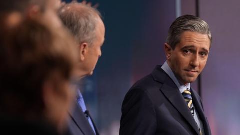 Taoiseach and leader of Fine Gael Simon Harris during the General Election leaders' debate at RTE studios in Dublin. He is to the right of the picture, staring at his opponents who are blurred. He is wearing a navy jacket and yellow and blue striped tie.