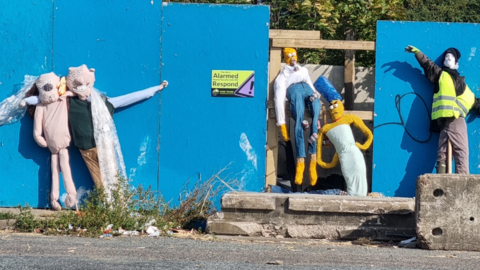 Scarecrows resembling Wallace and Grommet, Homer and Marge Simpson and a man in a high vis vest with a speed gun have been nailed to a blue wall 