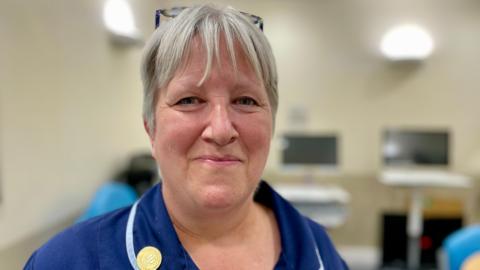 A nurse in a blue uniform with ash blonde hair smiling at the camera standing in a clinical room in a hospital  with computer screens in the background. 