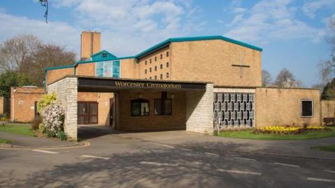 Worcester crematorium displaying the black sign above the entrance. The building has a teal roof and light brown bricks. There is a tall cross on the side of the building.