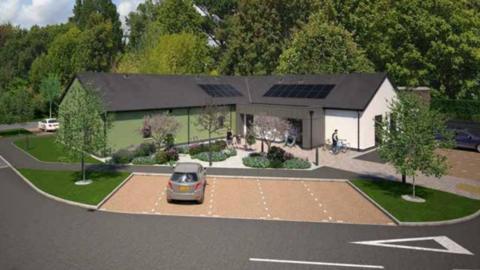 A CGI of the building which is a modern centre, and is green on one side and white on the other, there are solar panels on the sloped roof and trees around.