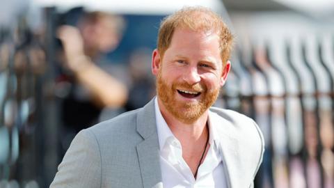 Prince Harry wearing a pale grey jacket and white shirt, smiling broadly - he is outside and a metal fence can be seen behind him