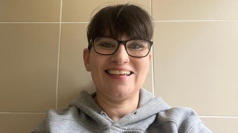 A woman with dark reading glasses and dark hair smiles into the camera. She is wearing a grey hooded top, and has a nose ring.