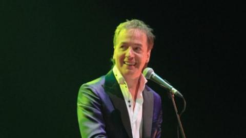 A man is illuminated by a green light. He is stood with a microphone in front of him and is looking sideways whilst smiling. He is wearing a black suit jacket and white shirt with collar and top buttons undone.