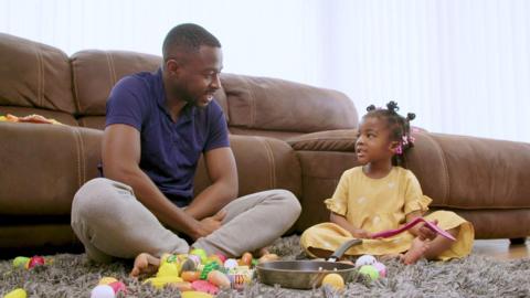 Father and daughter play with food toys and a frying pan in the living room.