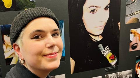 AJ Turner wearing a black beanie and has short blonde hair taking a picture with a an image of her younger self at the exhibition. 