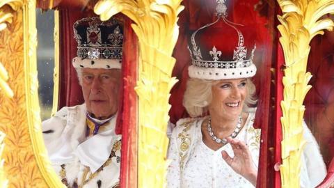 King Charles and Queen Camilla sat in a red Royal carriage detailed with gold leaves. They are both wearing white and gold, and each have an elaborate Royal crown on their heads.