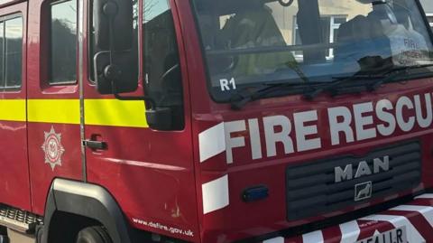 The picture shows a red fire engine with "FIRE RESCUE" displayed on the front.
A yellow stripe runs horizontally along the side of the truck.
The fire department logo is visible.
The website "www.dsfire.gov.uk" is printed near the bottom.