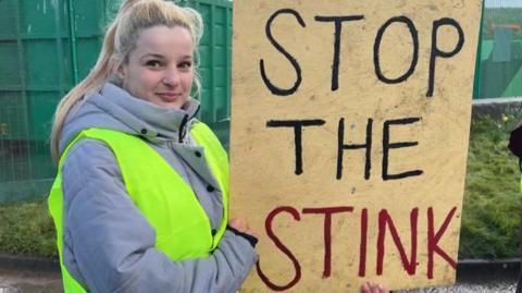 A young woman with long blonde hair, in a grey coat and yellow high vis vest, holds a sign that says 'stop the stink' in front of large green skips at a landfill site.