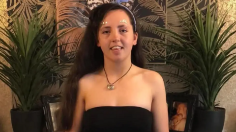 Maria pictured with long black thick hair, wearing a black top, a silver heart necklace and glitter stickers above her eyebrows. There are tow small palm like plants on either side of her.