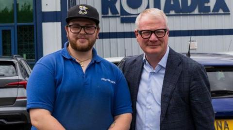 Daniel Harte with a blue top, a black cap, a beard and glasses is on the left next to Richard Parker, who is wearing a shirt, a dark jacket and glasses. Parked cars and a building are behind them.