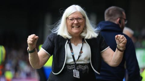 Port Vale Football Club owner Carol Shanahan is seen wearing glasses, a lanyard, white blouse and black jacket with white piping. She is clenching her fists and smiling in celebration.