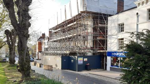 A hotel building covered in scaffolding and with a large barricade set up at the base. It is pictured between other buildings at the side of a street.