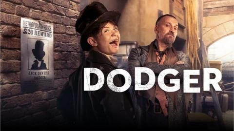 Dodger, a young boy is covered in soot and dirt in Victorian London, he is wearing a scruffy top hat and black coat and smiling, a wanted poster with his face on is on a wall behind him.