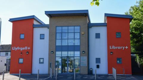 Prestatyn library building with orange painted walls either side bearing the words Llyfrgell+ and Library+ either side of a column of white and the glass-fronted entrance with bollards in front