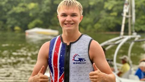 A boy with blonde hair is standing by the water giving both thumbs up. He is wearing a neoprene vest in the British flag colours.  